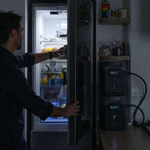 Ein Mann steht in einer schwach beleuchteten Küche und greift in einen offenen Kühlschrank. Das Innere des Kühlschranks ist hell erleuchtet und zeigt verschiedene Lebensmittel und Behälter, darunter Obst und Getränke. Auf der rechten Seite stehen auf einem Holzregal zwei elegante Geräte mit digitalen Anzeigen, auf denen die Temperatur eingestellt werden kann. Die Küche hat eine moderne Ästhetik, mit einer dezenten Farbpalette und ordentlich angeordneten Tassen und Gläsern in den Regalen darüber.