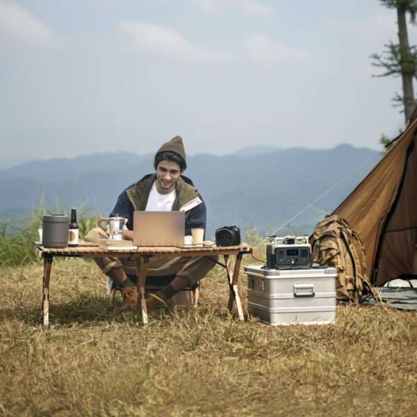 Ein Mann sitzt an einem Tisch auf einem Feld und arbeitet an einem Laptop, im Hintergrund ein Zelt, eine Bergkette, Himmel, sonniger Tag, warme Farbtöne, natürliches Licht, realistischer Stil, Abenteurer, Outdoor-Umgebung, digitaler Nomade, gemütliche Atmosphäre.