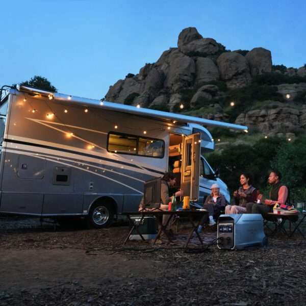 Eine gemütliche Campingszene zeigt ein graues, mit Lichterketten beleuchtetes Wohnmobil, das in der Nähe eines felsigen Hangs geparkt ist. Vier Personen sitzen auf Klappstühlen um einen kleinen Tisch herum, unterhalten sich und genießen Getränke. Die Atmosphäre ist entspannt, in der Nähe stehen eine Kühlbox und ein tragbares Kraftwerk. Der Himmel geht von der Dämmerung in die Abenddämmerung über, was die Atmosphäre im Freien noch heiterer macht.