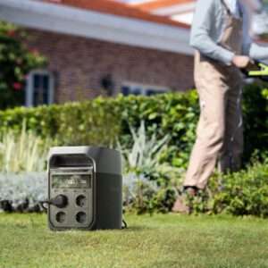 Ein kompaktes, tragbares Kraftwerk steht auf grünem Gras in einem Garten. Es verfügt über mehrere Steckdosen und eine digitale Anzeige für den Batteriestand. Im Hintergrund benutzt eine Person in legerer Arbeitskleidung ein Gartengerät, während sie von üppigem Grün und einer gepflegten Hecke umgeben ist. Ein Haus mit rotem Ziegeldach ist teilweise hinter dem Laub sichtbar.
