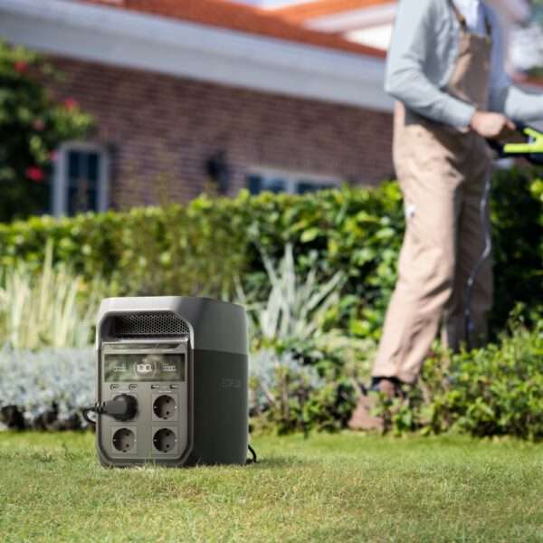 Ein kompaktes, tragbares Kraftwerk steht auf grünem Gras in einem Garten. Es verfügt über mehrere Steckdosen und eine digitale Anzeige für den Batteriestand. Im Hintergrund benutzt eine Person in legerer Arbeitskleidung ein Gartengerät, während sie von üppigem Grün und einer gepflegten Hecke umgeben ist. Ein Haus mit rotem Ziegeldach ist teilweise hinter dem Laub sichtbar.