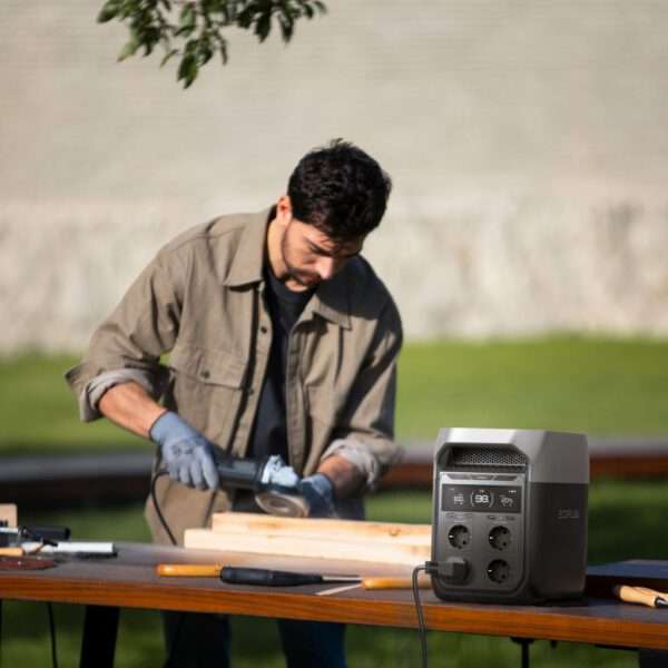 Ein Mann mit beiger Jacke und grauen Handschuhen arbeitet an einem Außenarbeitsplatz mit einem Elektrowerkzeug an einem Stück Holz. Im Vordergrund ist eine EcoFlow-Energiestation mit mehreren Steckdosen eingesteckt, die das Werkzeug mit Strom versorgt. Die Umgebung besteht aus einer grünen Wiese und einer Steinstruktur im Hintergrund, die auf einen hellen, sonnigen Tag hinweist.