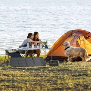 Das 45W portable Solarpanel von EcoFlow aufgestellt, daneben die River 3, die durch das Solarpanel geladen wird. Dahinter zwei Personen die an einem Campingtisch sitzen und an einem See mit ihrem Hund campen.