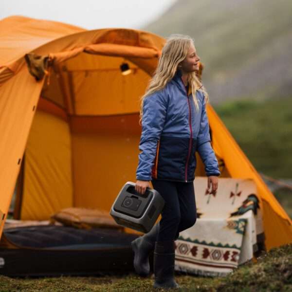 Ein junges Mädchen mit langen, welligen blonden Haaren lächelt glücklich, während sie im Freien in der Nähe eines leuchtend orangefarbenen Campingzeltes spazieren geht. Sie trägt eine blaue und marineblaue wasserdichte Jacke, eine dunkle Hose und schwarze Gummistiefel. In ihrer rechten Hand hält sie einen kompakten tragbaren Lautsprecher mit einem modernen, schlanken Design. Der Hintergrund zeigt eine üppige, grüne Landschaft mit sanften Hügeln und nebligen Bergen, die eine ruhige, natürliche Kulisse schaffen. Weiches, diffuses Licht suggeriert einen bewölkten Tag und verstärkt die gemütliche und abenteuerliche Atmosphäre. Die Szene fängt einen Moment der Freude und Entdeckung in einer wilden Campingumgebung ein, wobei die detaillierten Texturen des Zeltstoffs, der Haare des Mädchens und der natürlichen Umgebung zu einer warmen, einladenden Ästhetik beitragen. Der Stil ist realistisch, mit einem Fokus auf lebendigen Farben und natürlichem Licht, wodurch das Outdoor-Abenteuer und die jugendliche Begeisterung betont werden.