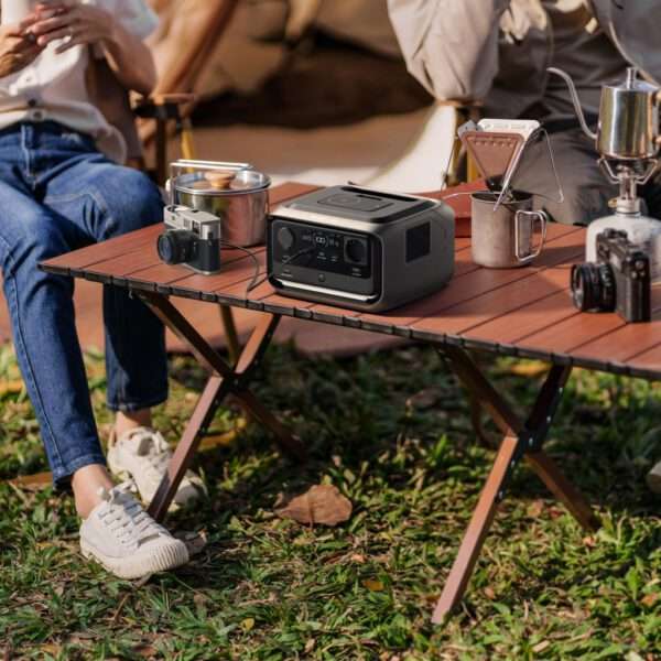 Nahaufnahme eines hölzernen Campingtisches, der im Freien auf Gras mit verstreuten Blättern steht und auf dem verschiedene Camping- und Fotoausrüstung zu sehen ist. Im Mittelpunkt steht ein moderner tragbarer Lautsprecher mit einem schlanken Design in Schwarz und Silber, einem digitalen Display und Bedienknöpfen. Links davon liegt eine Kamera im Vintage-Stil mit einem grauen Trageriemen auf dem Tisch, daneben ein kleiner Metalltopf mit Henkel. Rechts neben dem Lautsprecher steht ein Metallbecher mit Henkel, auf dem ein kleiner Klappkocher mit einem Wasserkocher steht, sowie ein kompakter tragbarer Kocher mit Metallic-Finish und einer kleinen Flamme. Ein Smartphone ist auf einem Ständer neben dem Kocher aufgestellt. Die Szene umfasst auch eine zweite Kamera mit einem größeren Objektiv, die ganz rechts positioniert ist, und auf der linken Seite des Bildes sind die Beine einer Person in lässigen Blue Jeans und weißen Turnschuhen zu sehen, deren Hände auf dem Schoß ruhen. Der Hintergrund suggeriert eine gemütliche Umgebung im Freien, möglicherweise einen Campingplatz oder einen Picknickplatz, mit warmem natürlichem Licht und einer entspannten Atmosphäre. Der Gesamtstil ist realistisch mit detaillierten Texturen der Ausrüstung, natürlichen Lichteffekten und einem Fokus auf Outdoor-Abenteuer und Fotoausrüstung.