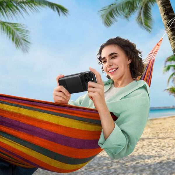 Eine junge Frau entspannt sich in einer bunt gestreiften Hängematte an einem Strand, hält ein Smartphone in der Hand und lächelt, während sie ihre Zeit genießt. Palmen rahmen die Szene ein, während der klare Himmel und der Sandstrand im Hintergrund eine sonnige, tropische Atmosphäre suggerieren.