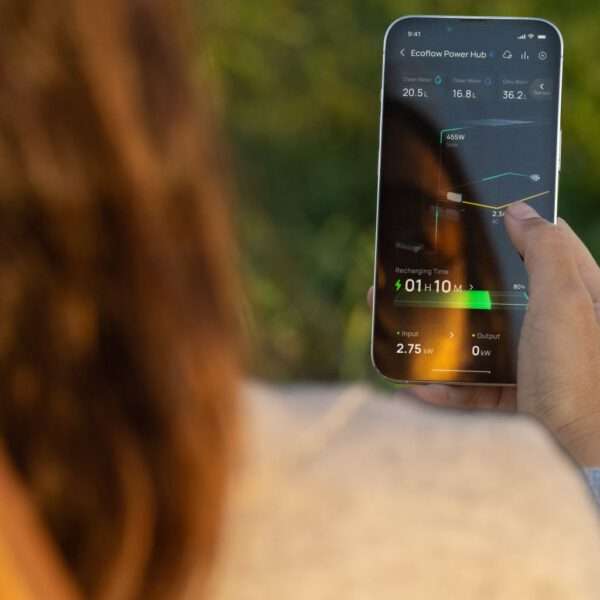 Eine Person mit langen, gewellten Haaren hält ein Smartphone in der Hand, auf dem die Oberfläche der Ecoflow Power Hub-App angezeigt wird. Der Bildschirm zeigt verschiedene Datenpunkte an, darunter die aktuelle Energiezufuhr (2,75 kW), die Leistung (0 kW) und die Wiederaufladezeit (01h 10m) sowie die Wasserstände. Der Hintergrund ist eine sanft verschwommene Grünfläche, die eine Außenumgebung suggeriert.