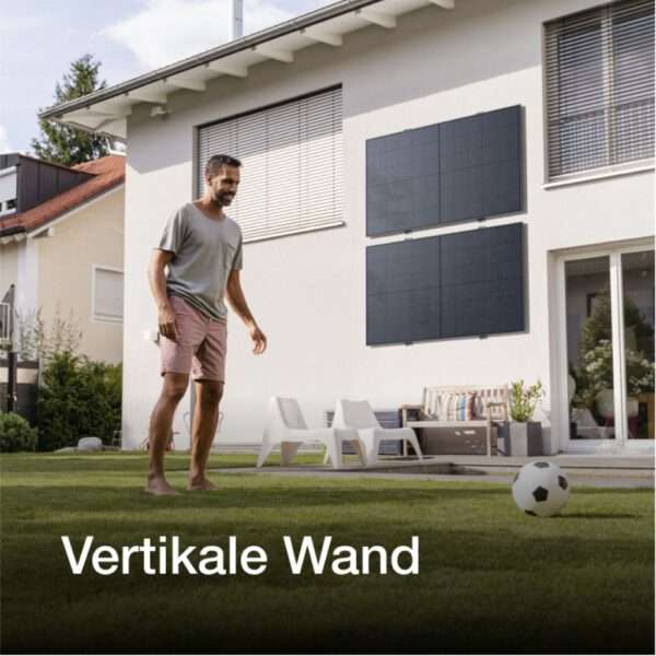 Ein Mann in legerer Kleidung steht auf einem gut gepflegten Rasen und bereitet sich darauf vor, einen Fußball zu kicken. Im Hintergrund befindet sich ein modernes Haus mit zwei an der Wand montierten Sonnenkollektoren und einem Sitzbereich mit zwei weißen Stühlen und einem kleinen Tisch. Die Umgebung ist hell und sonnig und spiegelt eine typische Szene im Freien in einem Wohngebiet wider.