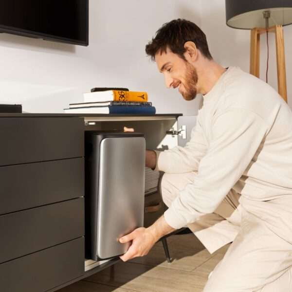 Ein Mann in heller Freizeitkleidung lächelt, während er neben einem modernen Fernsehschrank kauert. Er zieht einen eleganten, silbernen Mülleimer aus einem offenen Fach darunter hervor. Auf dem Schrank stapeln sich Bücher, darunter ein farbenfrohes Exemplar mit gelbem Buchrücken, das vom warmen Sonnenlicht aus einem nahe gelegenen Fenster beleuchtet wird. Im Hintergrund ist eine stilvolle Stehlampe zu sehen.