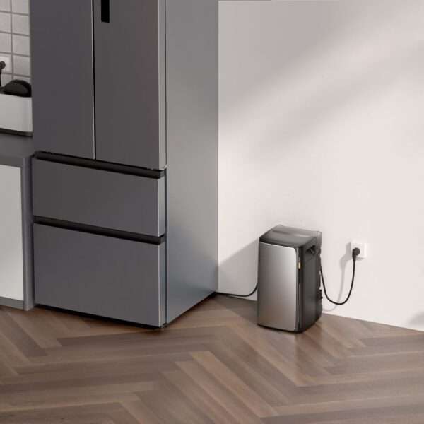 Eine moderne Küchenecke mit einem eleganten, dunkelgrauen Kühlschrank mit Gefrierfach unten, der neben einem grauen Kompaktgerät steht, möglicherweise einem kleinen Gefrierschrank oder einer Küchenmaschine, das an eine Steckdose an der Wand angeschlossen ist. Der Boden hat ein Fischgrätenmuster aus Holz, und der Hintergrund besteht aus einer weißen Wand und einer gefliesten Küchenrückwand. Natürliches Licht wirft sanfte Schatten in den Raum und unterstreicht die moderne Ästhetik.