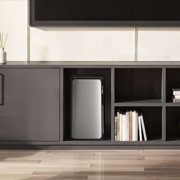 Ein modernes, minimalistisches Unterhaltungsmöbelstück besteht aus einem eleganten schwarzen Schrank mit offenen und geschlossenen Regalen. Ein kleiner silberner Kühlschrank ist ordentlich in eines der Fächer eingebaut und präsentiert seine glänzende Oberfläche. Auf dem angrenzenden Regal sorgen mehrere ordentlich angeordnete Bücher und ein dekorativer Glasgegenstand für einen Hauch von Stil. Den Hintergrund bildet ein großer Flachbildfernseher, der über dem Möbelstück angebracht ist, wobei eine helle Wand einen zeitgemäßen Akzent setzt. Der Holzboden reflektiert das natürliche Licht und unterstreicht die Gesamtästhetik.