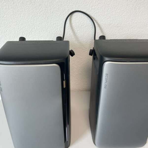 Zwei graue und schwarze tragbare EcoFlow-Kraftwerke sind nebeneinander auf einer weißen Fläche aufgestellt. Sie zeichnen sich durch ein schlankes Design mit metallischen Fronten und schwarzen Oberseiten aus. Ein dünnes schwarzes Kabel verbindet die beiden Geräte, an deren Seiten Stromtasten und Anschlüsse sichtbar sind. Der Hintergrund ist eine schlichte weiße Wand.