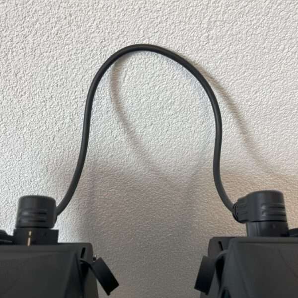 Eine Nahaufnahme eines schwarzen Kabels, das zwei elektronische Geräte auf einer strukturierten weißen Wand verbindet. Das Kabel ist in der Mitte gebogen, so dass eine Schleife entsteht, und die Enden sind in die Geräte eingesteckt, die ein einfaches, rechteckiges Design haben. Der Hintergrund ist eine schlichte, helle Wand, die den Kontrast zu dem schwarzen Kabel und den Geräten hervorhebt.