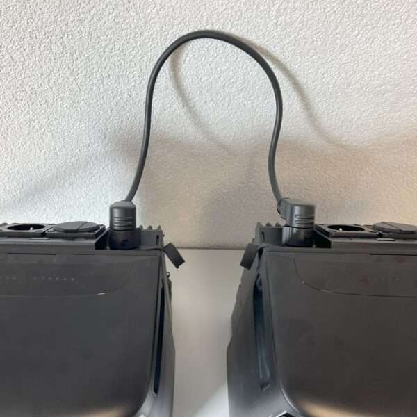 Zwei schwarze elektronische Geräte liegen dicht nebeneinander auf einer Fläche und sind oben mit einem einzigen schwarzen Kabel verbunden. Die Geräte scheinen glatte Oberflächen und nur wenige sichtbare Tasten oder Anschlüsse zu haben, mit Ausnahme einiger weniger auf der Oberseite. Als Hintergrund dient eine strukturierte weiße Wand.