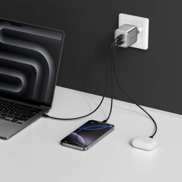 In einem eleganten, modernen Arbeitsbereich steht ein Laptop mit einem dunklen, abstrakten Bildschirm neben einem Smartphone und einem Paar kabelloser Ohrhörer. Die Geräte sind an ein kompaktes silbernes Ladegerät angeschlossen, das in einer Steckdose steckt und von dem mehrere schwarze Ladekabel abgehen. Der minimalistische Schreibtisch ist weiß und bildet einen klaren Hintergrund vor einer dunkelgrauen Wand.