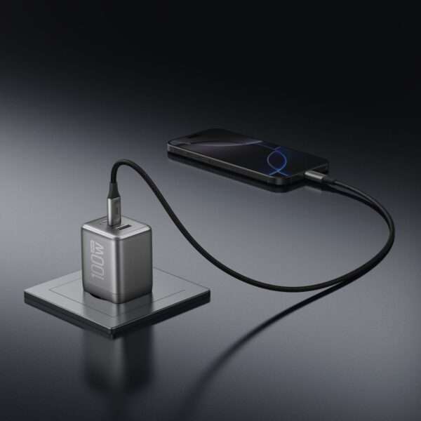 Ein schlankes, modernes Ladegerät mit der Bezeichnung 100 W ruht auf einer glänzend schwarzen Oberfläche und ist über ein geflochtenes Kabel mit einem Smartphone verbunden. Das Telefon hat ein minimalistisches Design, das sein schlankes Profil hervorhebt. Der Hintergrund ist dunkel und hebt die polierten Metallstrukturen des Ladegeräts und des Telefons hervor, wodurch eine anspruchsvolle technische Ästhetik entsteht.