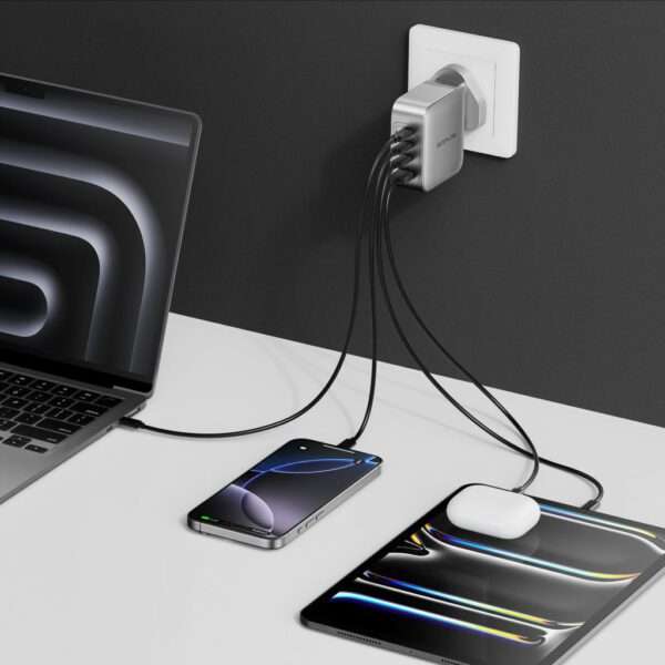 Ein eleganter, moderner Arbeitsplatz mit einem Laptop, der an ein an der Wand montiertes Ladegerät mit mehreren Anschlüssen angeschlossen ist. Das Ladegerät ist in eine Steckdose eingesteckt und verfügt über mehrere Kabel. Ein Smartphone und ein Tablet, beide mit glänzender Oberfläche, werden auf einer weißen Tischoberfläche aufgeladen. Das Smartphone zeigt eine grüne Akkuanzeige an, während auf dem Tablet ein Paar kabellose Ohrhörer liegt. Der Hintergrund ist ein dunkles Grau, das einen Kontrast zu den Geräten bildet.