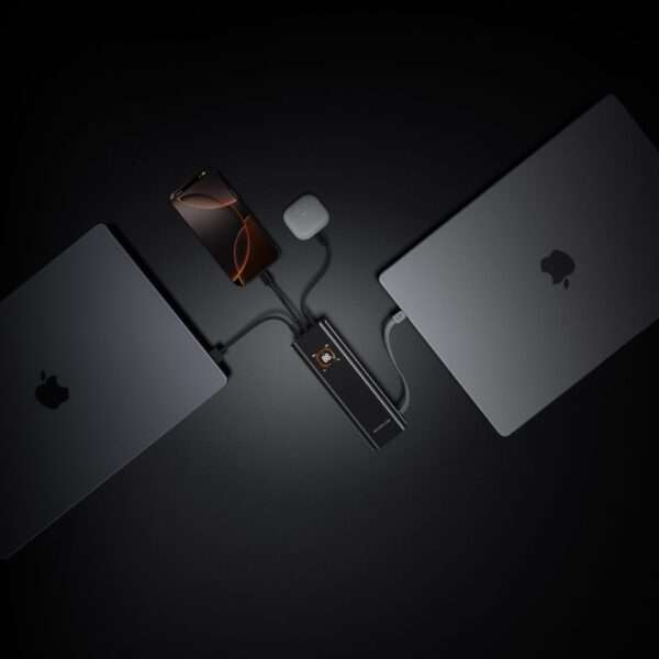 Eine elegante, minimalistische Einrichtung mit zwei MacBook-Laptops und einem Smartphone, die alle an einen kompakten Lade-Hub mit mehreren Anschlüssen angeschlossen sind. Die Ladestation verfügt über ein digitales Display und ist zentral positioniert, wobei die Kabel bis zu den Geräten reichen. Der Hintergrund ist dunkel und unterstreicht die moderne Ästhetik der technischen Geräte. Der Bildschirm des Smartphones zeigt ein dynamisches Hintergrundbild, das der monochromen Szene einen Farbakzent verleiht.