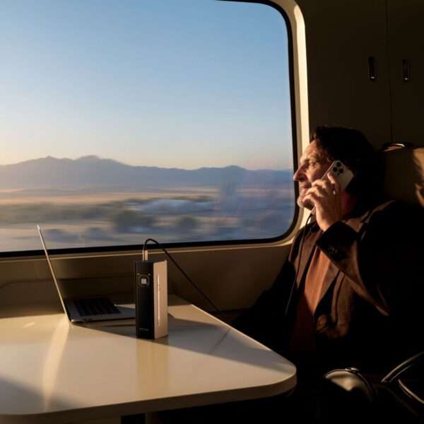 Ein Mann sitzt an einem Tisch in einem Zugabteil, telefoniert und arbeitet an einem Laptop. Im Hintergrund sieht man durch ein großes Fenster eine malerische Aussicht auf Berge und eine offene Landschaft, die von einem sanften Abendlicht erhellt wird. Auf dem Tisch liegt ein tragbares Gerät mit digitaler Anzeige, das mit dem Laptop verbunden ist. Der Ausdruck des Mannes ist konzentriert und in sein Gespräch vertieft.