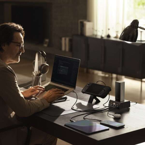 Ein Mann sitzt an einem modernen Schreibtisch und arbeitet an einem Laptop, auf dessen Bildschirm eine Programmier-Software angezeigt wird. Er hat eine Brille und graue Haare und trägt einen hellen Blazer. In der Nähe steht eine Spielkonsole auf einem Ständer, daneben befinden sich verschiedene Geräte wie ein Tablet, ein Smartphone und ein Paar Kopfhörer. Mehrere Ladekabel verbinden die Geräte mit einer eleganten Steckdose, während eine Schreibtischlampe den Arbeitsbereich beleuchtet. Im Hintergrund befindet sich ein minimalistischer Wohnbereich mit einem großen Fenster, das natürliches Licht hereinlässt.