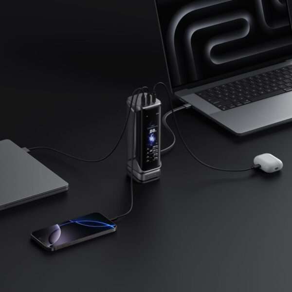 Eine elegante Powerbank in Metallic-Silber steht in der Mitte des Raums und ist über mehrere Kabel mit einem Laptop und einem Smartphone verbunden. Die Powerbank verfügt über ein digitales Display, auf dem Leistungsstatistiken angezeigt werden, während der Laptop und das Smartphone beide in dunklen Farben gehalten sind und das Smartphone einen leuchtenden Bildschirm hat. Ein Paar kabellose Ohrhörer ist ebenfalls angeschlossen und liegt in der Nähe. Der Hintergrund ist glatt und schwarz, was eine moderne und minimalistische Ästhetik schafft.