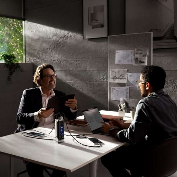 In einem lichtdurchfluteten, modernen Büro sitzen zwei Männer an einem weißen Tisch und unterhalten sich. Der eine Mann lächelt, hält ein Tablet in der Hand und trägt einen schwarzen Blazer. Der andere, der ein graues Hemd und eine Brille trägt, ist auf einen Laptop mit einem geöffneten Dokument konzentriert. Zwischen ihnen steht eine Ladestation für mehrere Geräte, an die mehrere Telefone angeschlossen sind. An der Wand im Hintergrund hängen Architekturzeichnungen, und durch ein Fenster fällt Tageslicht herein, das das moderne Design des Raums unterstreicht.