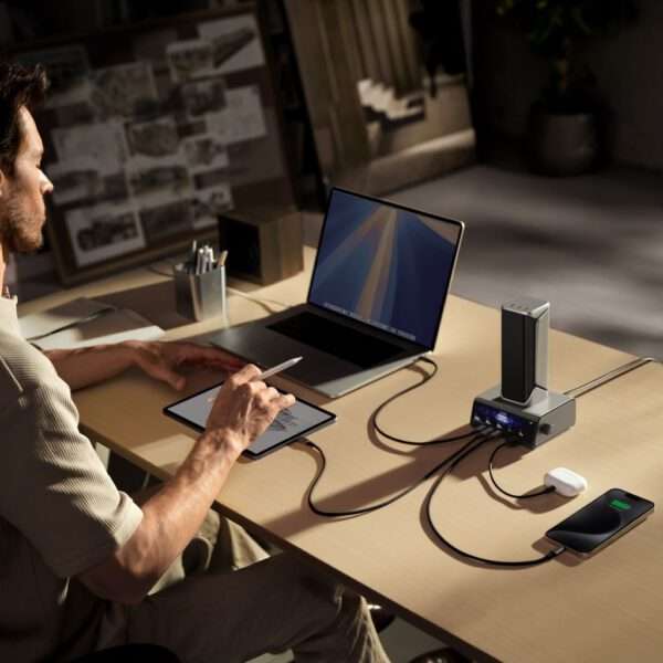 Ein Mann sitzt an einem Holzschreibtisch und arbeitet mit einem Stift in der Hand an einem Tablet. Vor ihm steht ein Laptop mit einem leuchtenden Bildschirm. Neben dem Laptop ist eine elegante Ladestation zu sehen, die über verschiedene Kabel mit einem Smartphone und einem Paar kabelloser Ohrhörer verbunden ist. Der Arbeitsbereich ist gut beleuchtet, mit sanften Schatten an der Wand, und im Hintergrund stehen Topfpflanzen und Skizzen, die eine moderne, aufgeräumte Atmosphäre schaffen.