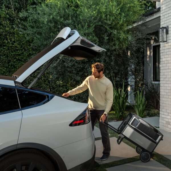 Ein Mann in einem hellen Pullover steht bei geöffneter Heckklappe neben einem schlanken, weißen Elektrofahrzeug. Er zieht eine Power Station mit Batterie auf Rädern zum Auto, und üppiges Grün umgibt die Szene, was die Atmosphäre im Freien noch verstärkt. Die Kulisse besteht aus einem modernen Haus mit klaren Linien und einer Backsteinfassade, die den zeitgenössischen Look des Augenblicks unterstreicht.