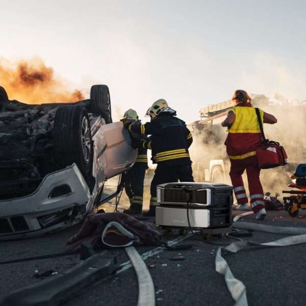 Eine dramatische Szene zeigt Rettungskräfte, die an der Unfallstelle eines umgestürzten Fahrzeugs Hilfe leisten. Zwei Feuerwehrleute mit Helmen und Schutzkleidung versuchen, eine Tür des umgestürzten Fahrzeugs zu öffnen, während ein dritter Helfer in einer rot-gelben Uniform in der Nähe steht und bereit ist zu helfen. Rauch und Flammen wallen im Hintergrund und machen die Rettungsarbeiten noch dringlicher. Auf dem Boden liegen verstreute Ausrüstungsgegenstände, darunter Schläuche und ein tragbares Kraftwerk, die das Chaos der Situation verdeutlichen. Der Sonnenuntergang wirft ein warmes Licht auf die Szene.