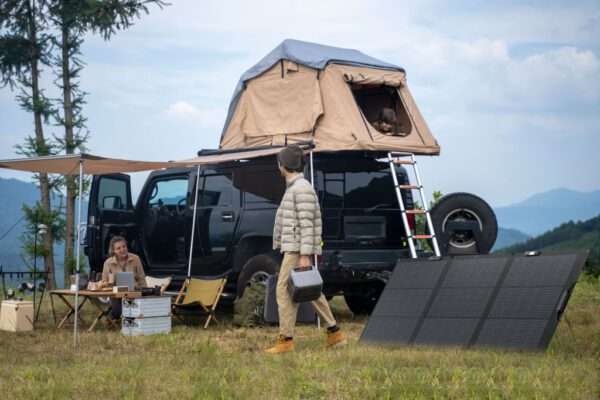 A rugged black SUV with a rooftop tent is parked on a grassy area surrounded by trees and mountains. A woman is seated at an outdoor table working on a laptop, while a man, dressed in a light puffer jacket and carrying a portable power station, walks towards her. A solar panel setup rests on the ground nearby, and a child is visible peeking out from the rooftop tent's opening. The sky is partly cloudy, adding to the scenic outdoor atmosphere.