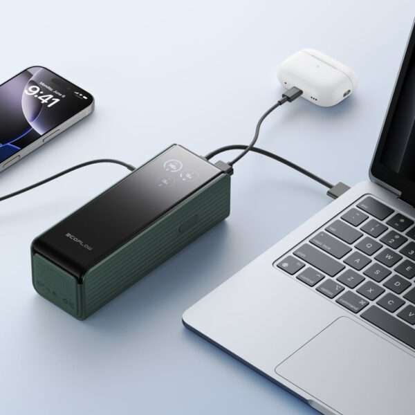 Auf einer hellblauen Fläche steht eine grüne, tragbare Powerbank mit glatter schwarzer Oberseite und digitalem Display, die über ein schwarzes Kabel mit einem Paar weißer, kabelloser Ohrhörer verbunden ist. In der Nähe befindet sich ein silberner Laptop mit schwarzer Tastatur, der teilweise sichtbar ist und über einen USB-C-Anschluss verfügt, der ebenfalls mit der Powerbank verbunden ist. Links neben der Powerbank steht ein iPhone, das die Uhrzeit von 9:41 Uhr anzeigt.