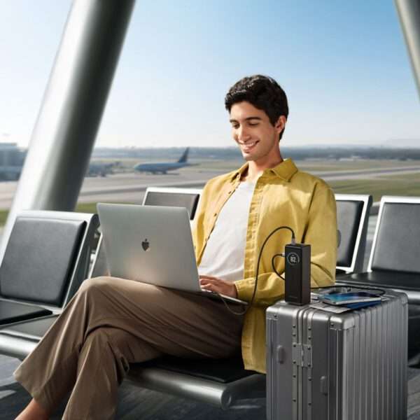 Ein junger Mann sitzt bequem in einem Flughafenterminal und arbeitet lächelnd an einem silbernen Laptop. Er trägt eine hellgelbe Jacke und eine beige Hose. Neben ihm stehen ein eleganter grauer Koffer und ein tragbares Ladegerät, auf dem der Akkustand angezeigt wird. Im Hintergrund ist eine Flughafenlandebahn zu sehen, auf der ein Flugzeug startbereit ist. Natürliches Licht erfüllt den Raum und unterstreicht die moderne Umgebung.