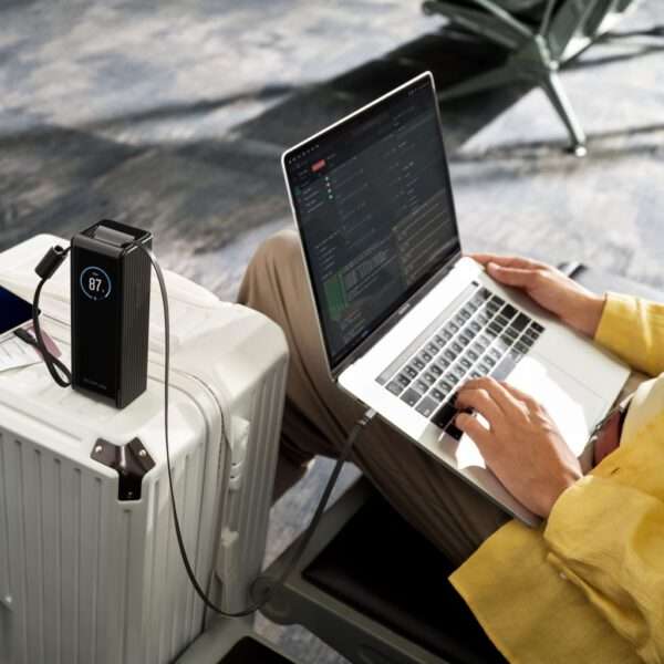Eine Person sitzt in einer ungezwungenen Umgebung und benutzt einen Laptop auf ihrem Schoß, während sie sich auf einen tragbaren weißen Koffer stützt. Ein schlankes schwarzes Gerät zeigt einen Zahlenwert von 87,7 an und ist über ein Kabel mit dem Laptop verbunden. Die Person trägt ein hellgelbes Hemd und scheint sich auf den Bildschirm zu konzentrieren, auf dem Zeilen mit Codes oder Daten angezeigt werden. Der Hintergrund weist eine weiche Textur auf, die zur Atmosphäre eines modernen Arbeitsbereichs beiträgt.