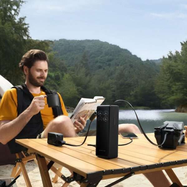 Ein Mann sitzt an einem Holztisch an einem ruhigen Seeufer und liest ein Buch, während er eine Tasse Kaffee genießt. Er ist leger mit einem gelben Hemd und einer schwarzen Weste gekleidet. Neben ihm ist ein tragbares Kraftwerk an eine Drohne und ein Smartphone angeschlossen, was auf eine entspannte Umgebung im Freien hindeutet. Üppig grüne Berge und Bäume bilden eine malerische Kulisse.