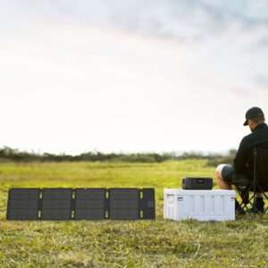 Eine Person sitzt auf einem Klappstuhl auf einer Wiese und blickt von der Kamera weg. Zu ihrer Linken ist eine tragbare Solaranlage ausgeklappt, die das Sonnenlicht einfängt. Daneben steht ein weißes Kraftwerk auf dem Boden, das mit den Solarzellen verbunden ist. Im Hintergrund ist eine ruhige Landschaft unter einem weichen, bewölkten Himmel zu sehen.