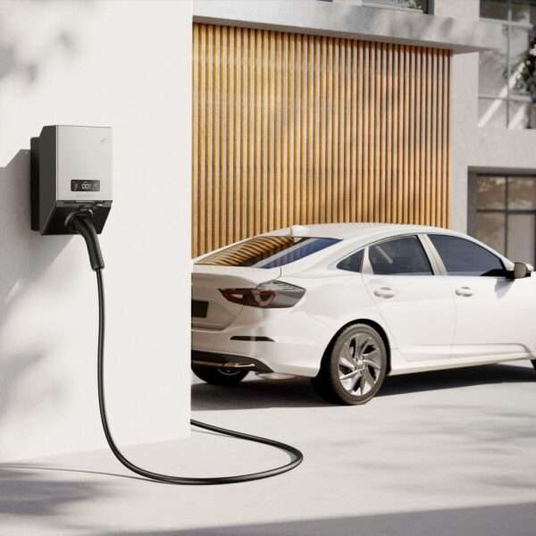 An einer Wand ist ein elegantes, modernes Ladegerät für Elektrofahrzeuge angebracht, aus dem ein Ladekabel herausragt. Im Hintergrund steht ein weißer Sedan in der Nähe einer Holzlattenwand, was auf eine moderne Wohnumgebung hindeutet. Die Szene ist gut ausgeleuchtet, wodurch klare Linien und eine minimalistische Ästhetik hervorgehoben werden.