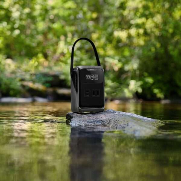 Eine tragbare Stromstation steht auf einem glatten Felsen in einem ruhigen Gewässer, umgeben von üppigem Grün. Das Gerät verfügt über ein digitales Display, das einen Prozentsatz anzeigt, und mehrere Ausgangsanschlüsse an der Vorderseite. Die Umgebung vermittelt eine friedliche Atmosphäre im Freien und spiegelt die Schönheit der Natur wider.