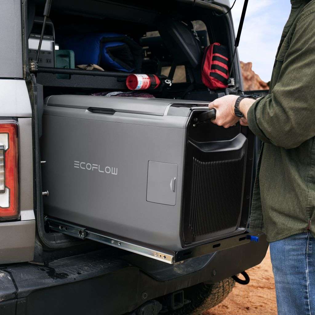 Eine Person zieht eine tragbare graue Kühlbox mit der Aufschrift ECOFLOW aus dem Kofferraum eines SUVs. Die Kühlbox steht auf einer Schiebeplatte, der Innenraum des Fahrzeugs ist zu sehen und enthält Campingausrüstung und einen roten Rucksack. Die Umgebung scheint im Freien zu sein, im Hintergrund sind rote Felsformationen zu sehen. Die Person trägt eine grüne Jacke und eine Uhr, was auf einen aktiven Outdoor-Lebensstil hindeutet.