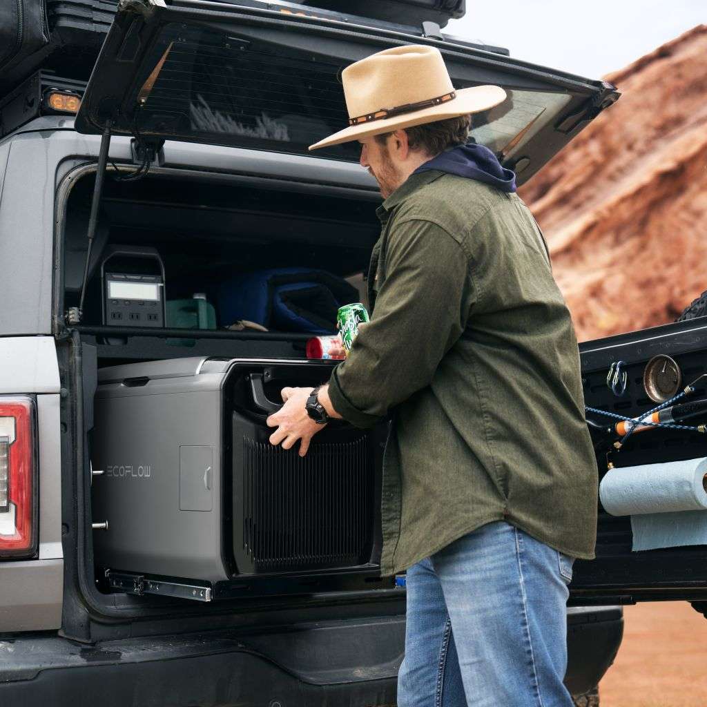 Ein Mann mit einem breitkrempigen Hut lädt einen EcoFlow-Campingkühlschrank in den Kofferraum eines grauen SUV. Er trägt ein grünes Hemd und Blue Jeans und hält in der einen Hand eine Dose Limonade, während er mit der anderen Hand den Kühlschrank feststellt. Im Hintergrund sind farbenfrohe Felsformationen zu sehen, und im SUV sind Outdoor-Ausrüstung und Vorräte zu erkennen.