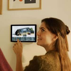 Eine Frau mit langen Haaren und einem warmen Lächeln interagiert mit einem an der Wand montierten digitalen Bedienfeld. Das Bedienfeld zeigt ein 3D-Modell eines Hauses mit Sonnenkollektoren sowie verschiedene Statistiken zum Energieverbrauch und zur Energieerzeugung an. Im Hintergrund verleihen gerahmte Kunstwerke dem Raum eine dekorative Note. Die Frau zeigt auf den Bildschirm und ist interessiert an den dargestellten Informationen.